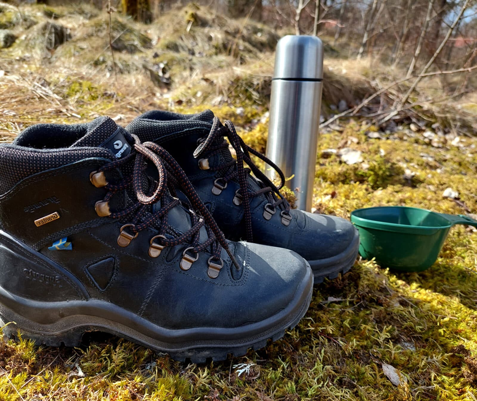 Kängor, termos och en kåsa som står på skogsmark